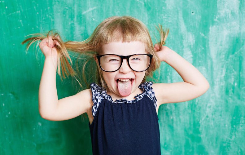 little blonde girl in pigtails and glasses pulling hair and sticking out tongue in front of chalkboard