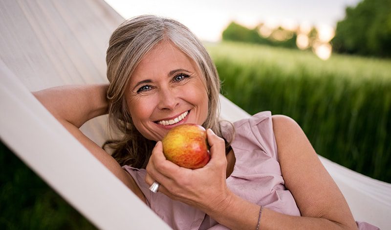 woman with sleeveless pink blouse, smiling, holding apple, and laying in hammock